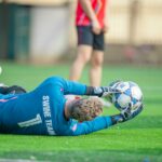 A goalkeeper in blue uniform catching a ball during a practice session on a green soccer field.
